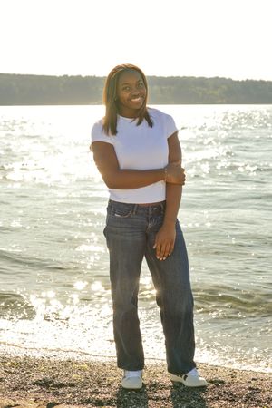 Smiling young woman in white t-shirt and jeans standing on a sunny beach by a sparkling lake.