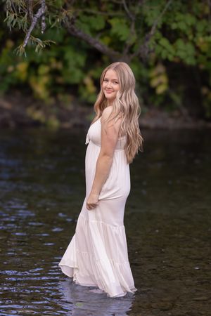 A person in a flowing white dress stands playfully in shallow water surrounded by trees and nature.