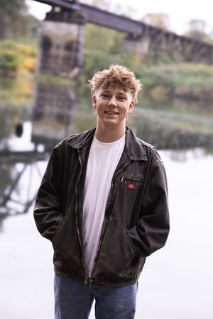 A young person in a black jacket and white t-shirt poses outdoors near a lake on an autumn day.