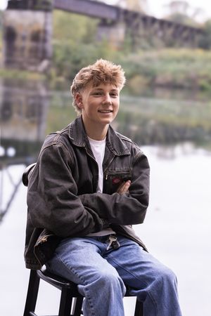 A person sits casually on a wooden bench wearing jeans and a dark jacket against a waterfront backdrop.