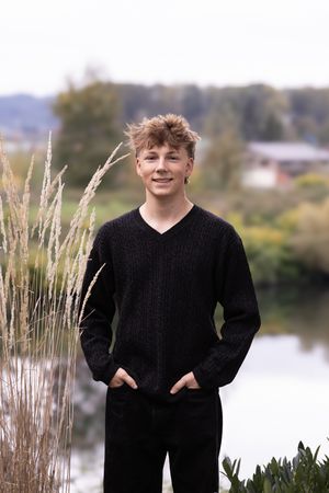 Person wearing a black sweater poses outdoors near a lake with tall grass and trees in the background.