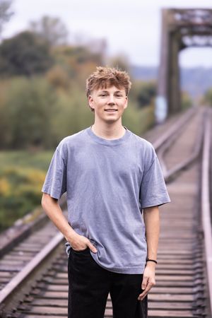 A young person stands on railroad tracks wearing a gray t-shirt and black pants on a cloudy day.