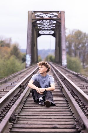 A person sits on railroad tracks leading to a steel bridge structure surrounded by autumn trees.