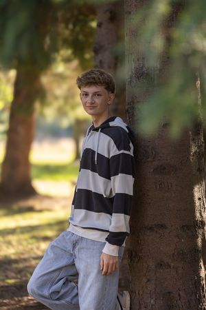 A person wearing a black and white striped sweater sits casually against a tree trunk in a sunlit outdoor setting.