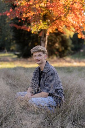 A person sits in a field of tall grass during autumn with colorful orange and yellow foliage in the background.