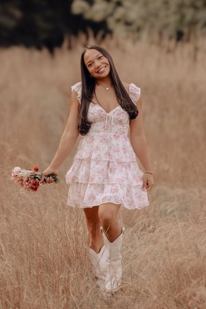 Young girl in white floral dress and boots runs playfully through golden grass field at sunset.