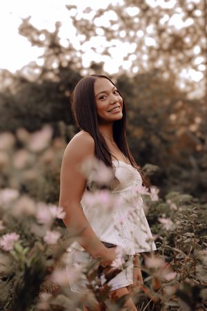 A woman in a white dress poses gracefully among blooming flowers during a golden hour photoshoot in nature.