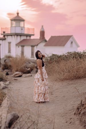 A white lighthouse stands against a pink sunset sky as a person in a floral dress explores the coastal landscape.