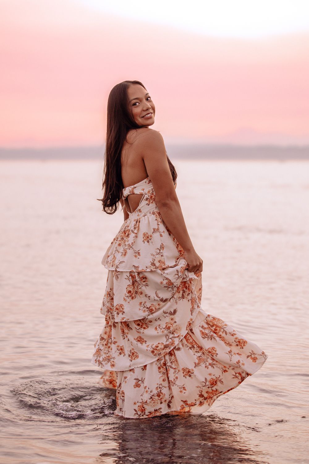 A woman in a flowing floral dress twirls joyfully in shallow ocean water during a pink sunset.