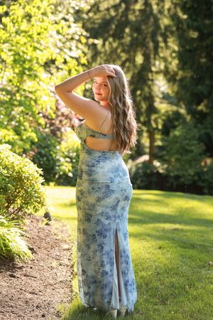 A person in a blue floral dress poses gracefully in a sunlit garden setting with lush green grass and trees.