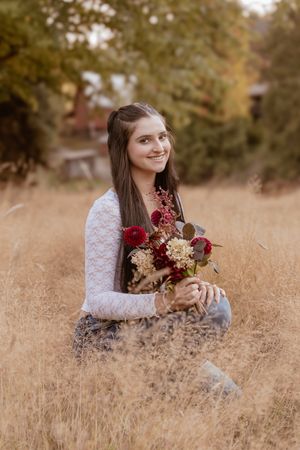 A person sits in a golden autumn field holding a bouquet of deep red and burgundy dried flowers.