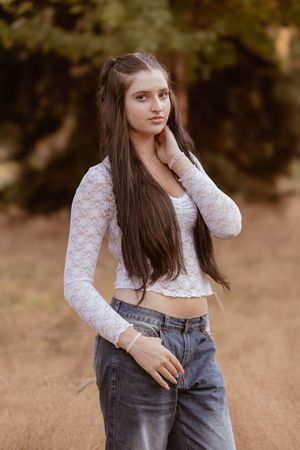 Young woman in white lace crop top and jeans poses outdoors against blurred natural background.