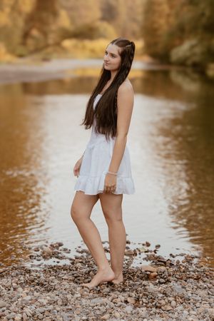 Person in white dress stands barefoot on rocky riverbank with autumn trees reflecting in calm water.