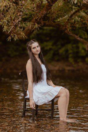 A white dress-clad figure sits on a chair in shallow creek water surrounded by autumn foliage.