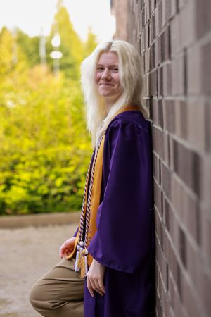 Woman in purple blazer and khaki pants poses against gray wall with autumn foliage in background.
