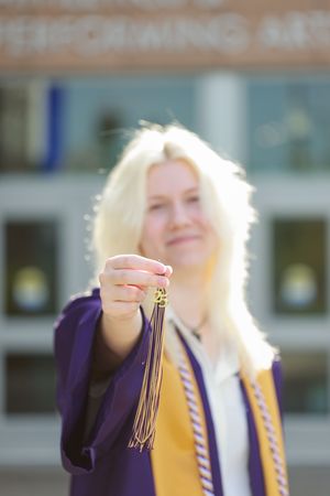 A person in a purple and gold graduation gown holds up their tassel while posing outdoors.