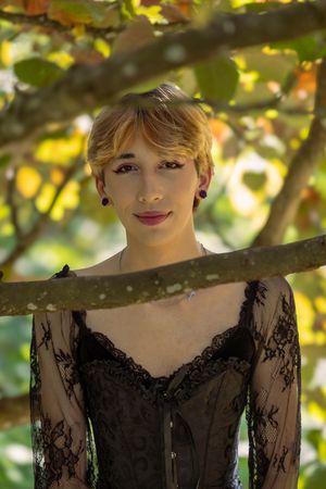 A figure in a black lace dress poses outdoors against a sunlit backdrop of green foliage and leaves.