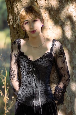 A figure in a black lace dress poses against a tree trunk in dappled sunlight creating a romantic gothic aesthetic.