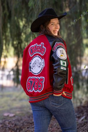 A person wearing a red varsity jacket and jeans poses in a wooded outdoor setting with blurred background.