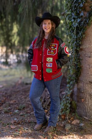 A person in a red varsity jacket and blue jeans poses playfully near trees in an outdoor setting.