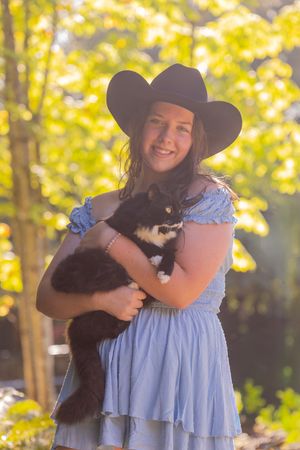 A person in a blue dress and sun hat holds a black cat outdoors with golden autumn foliage in the background.