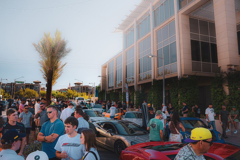People gather outside a modern office building for a car show featuring classic automobiles and sports cars.