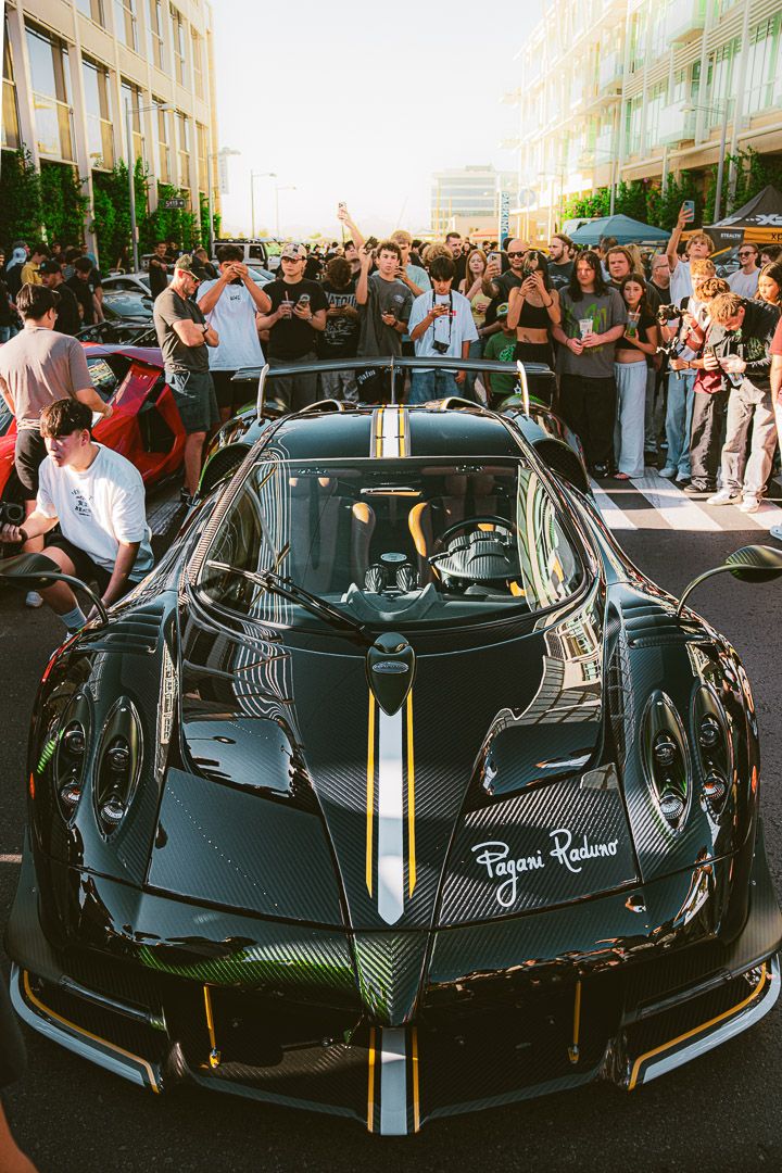 Black Pagani supercar with racing stripes displayed at a crowded automotive event.