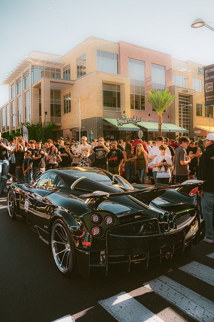 Crowd gathers around sleek black Ferrari supercars parked outside modern buildings during sunset event.