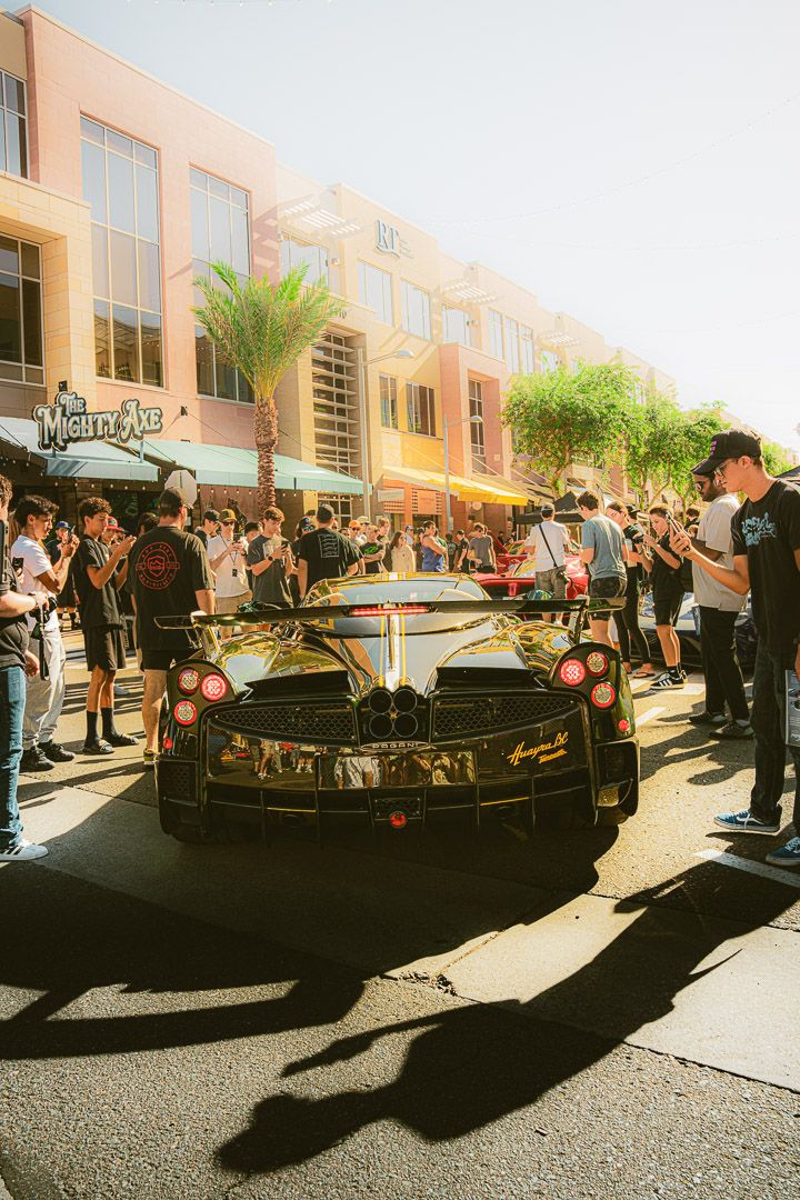 A sleek black sports car surrounded by spectators at a daytime street event with palm trees and buildings in the background.
