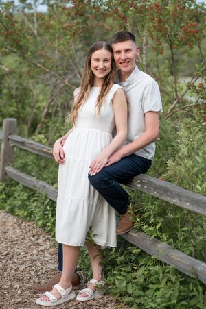 A couple shares an intimate moment sitting on a wooden fence surrounded by lush greenery and red berries.