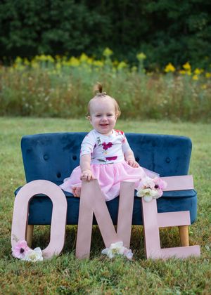 Baby in pink dress sits among large white 'ONE' letter props on blue chairs during outdoor first birthday photoshoot.