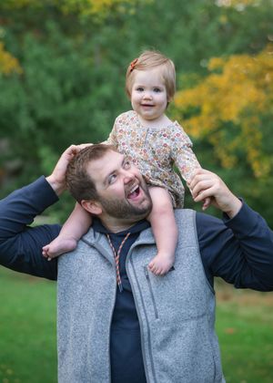 A joyful moment outdoors with a toddler riding on shoulders against a backdrop of yellow autumn flowers and greenery.