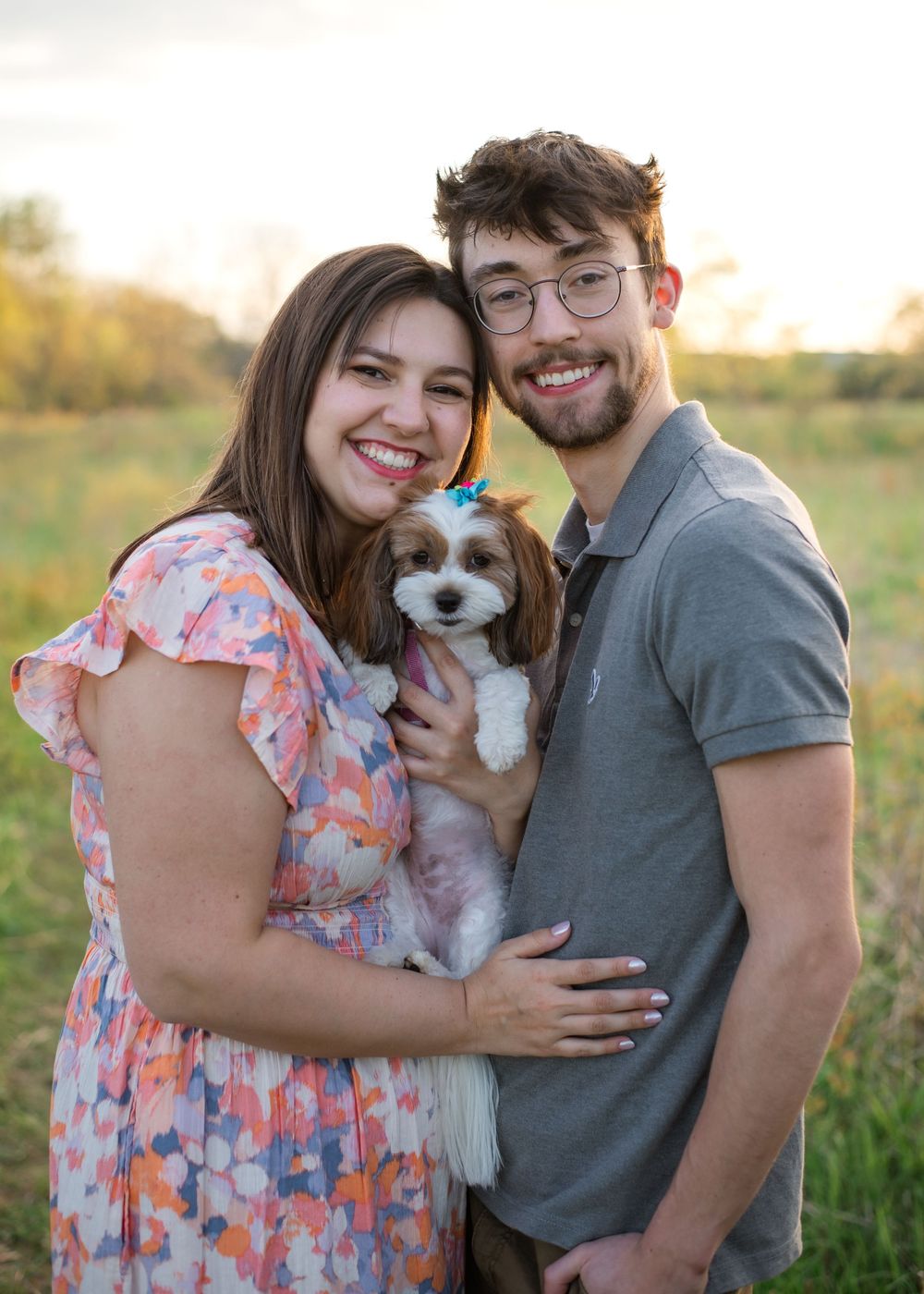 A couple holding their small brown and white dog in a field during golden hour while sharing affectionate moments.