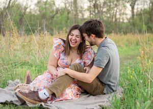 A couple sits together on a rustic wooden bench surrounded by tall grass in a summer meadow.