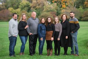 Large family group stands in a line on green grass with trees and autumn colors in the background.