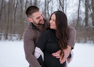 A romantic winter engagement photoshoot shows a couple embracing and laughing together in the snow-covered woods.