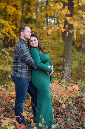 A couple embraces in an autumn forest setting with golden leaves, the woman wearing a long green maternity dress.