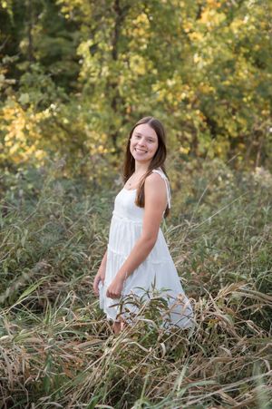 A person in a white dress poses in a field of tall grass with golden autumn foliage in the background.