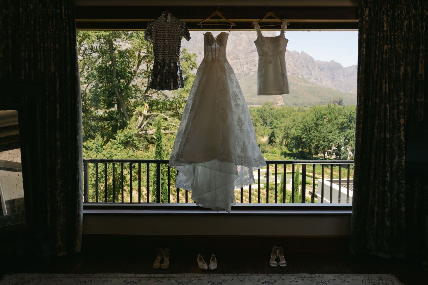 Three wedding dresses hanging in a window frame with mountain scenery and greenery visible in the background.