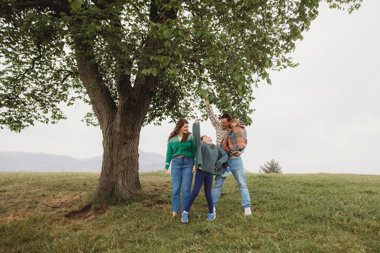 Three people stand together under a large tree on a grassy hilltop against an overcast sky.