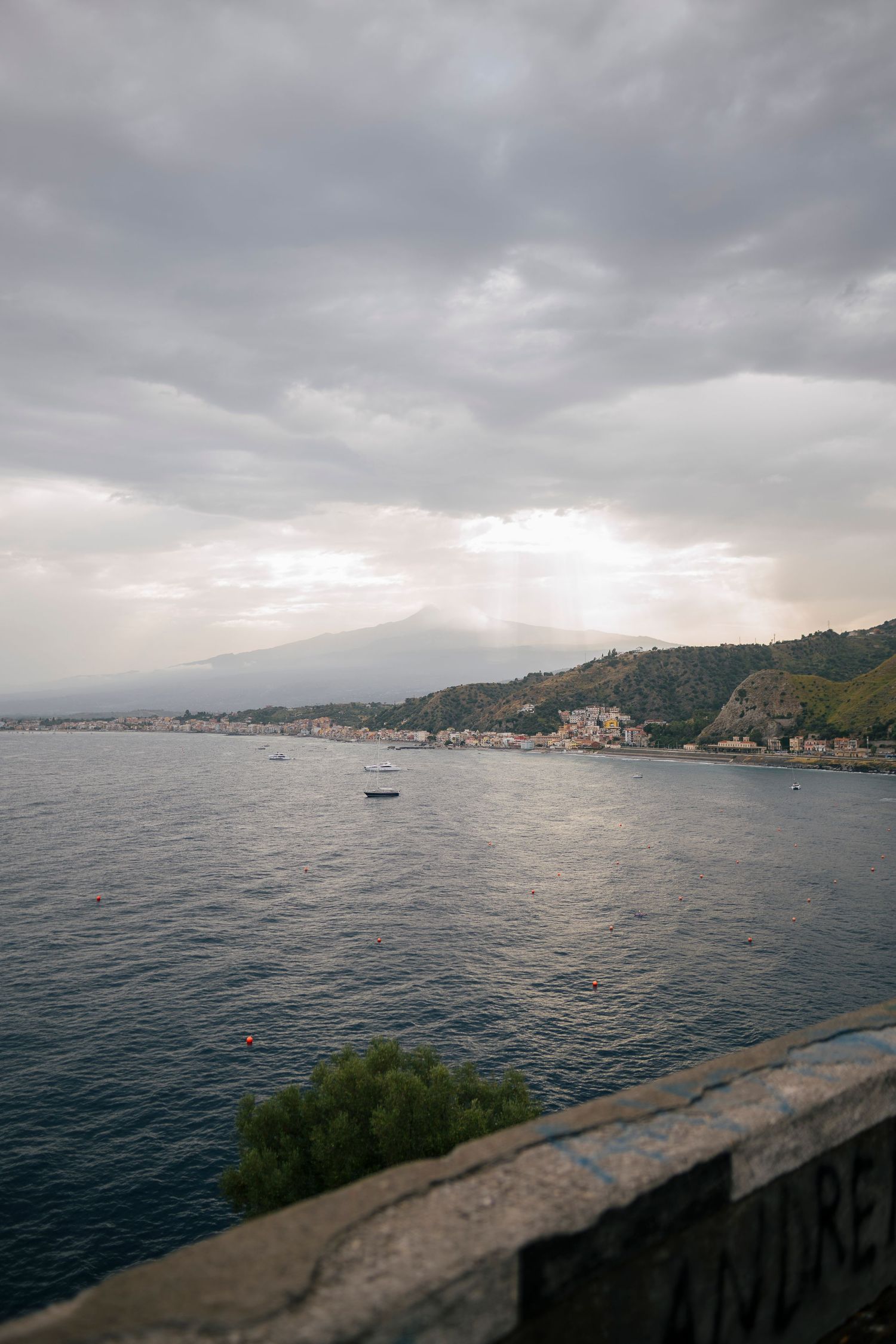 Dramatic cloudy sky over coastal Mediterranean waters with sunbeams breaking through clouds above hillside buildings.
