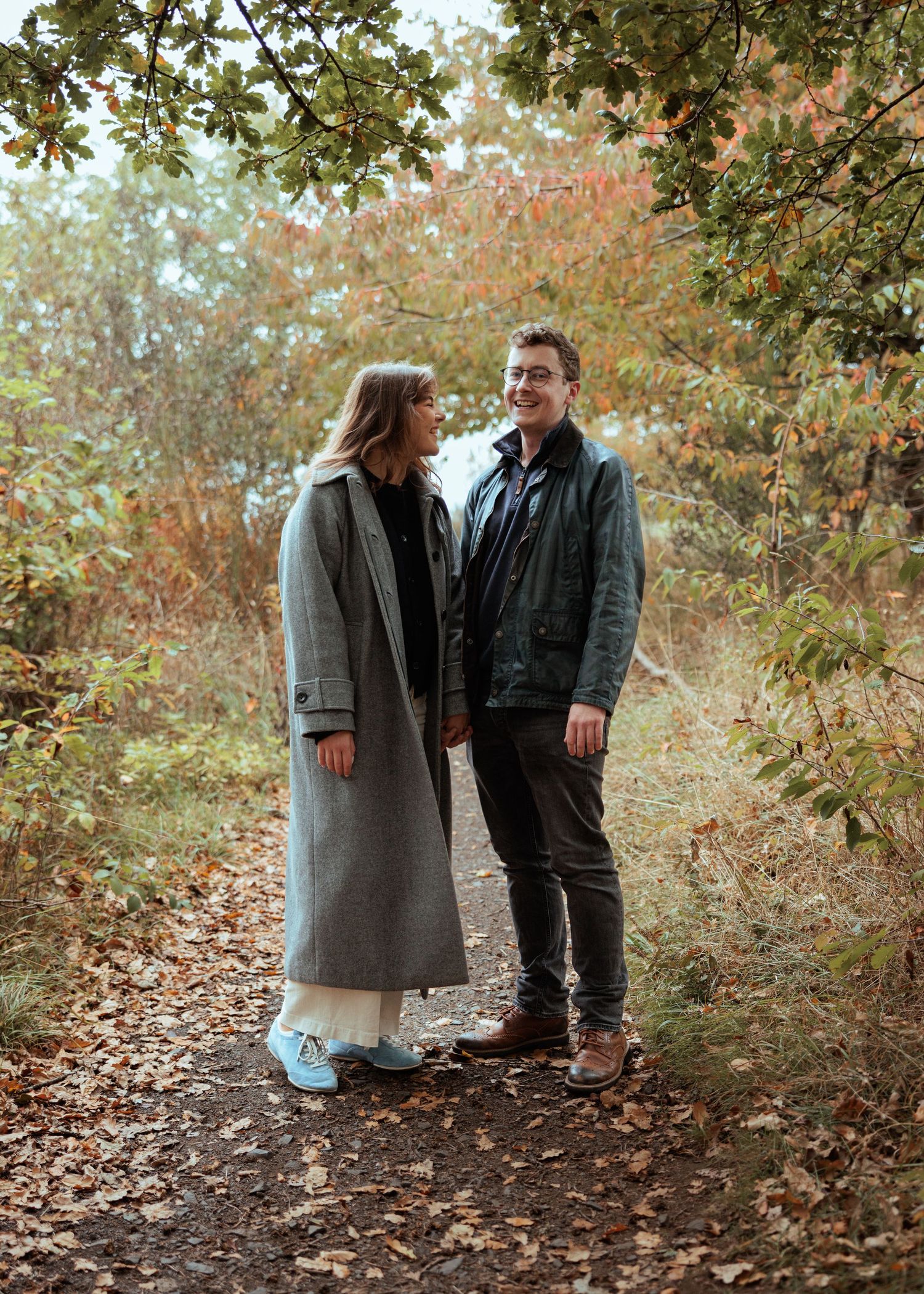 A couple in casual fall attire stands together on a leaf-covered forest path during autumn.