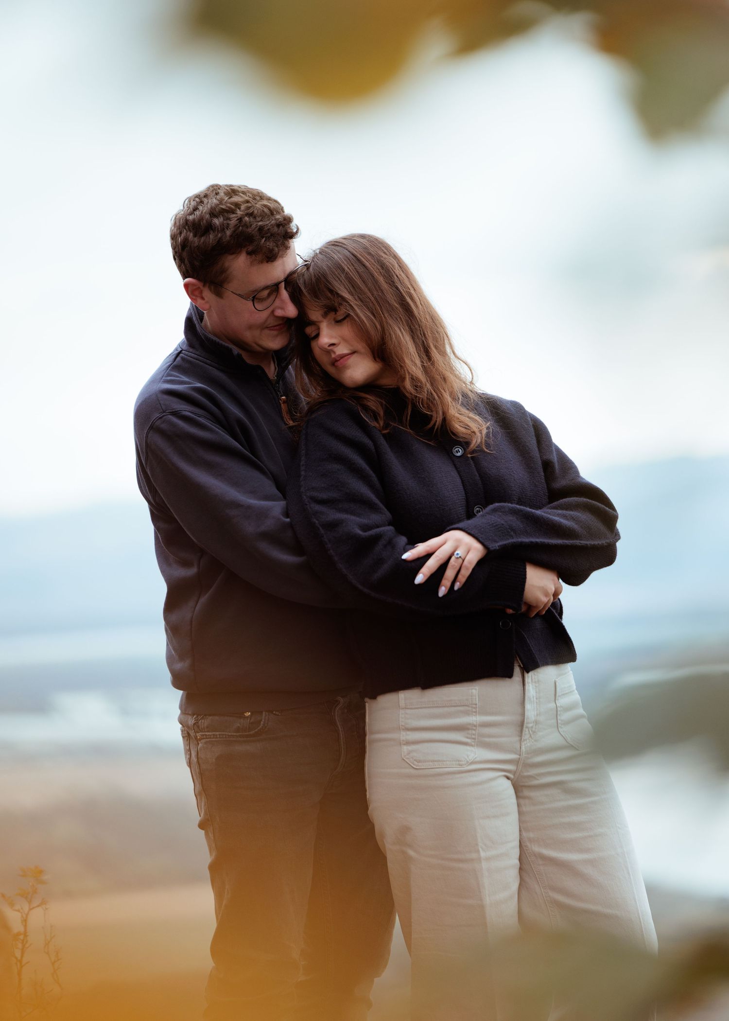 A romantic couple in matching dark jackets embrace tenderly against a soft blurred background.