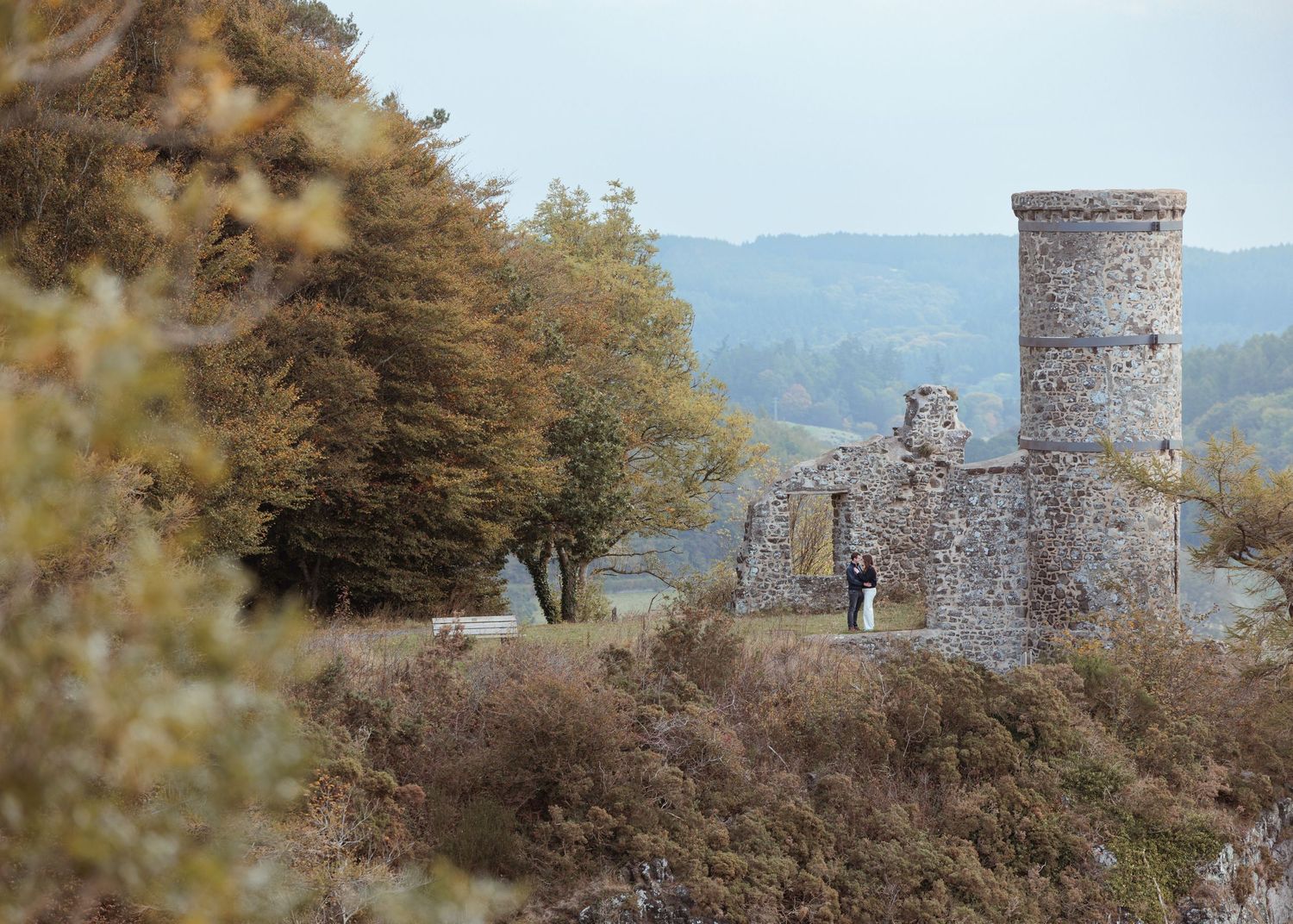 Ancient stone tower stands atop a windswept hill surrounded by autumn foliage and distant mountains in the background.