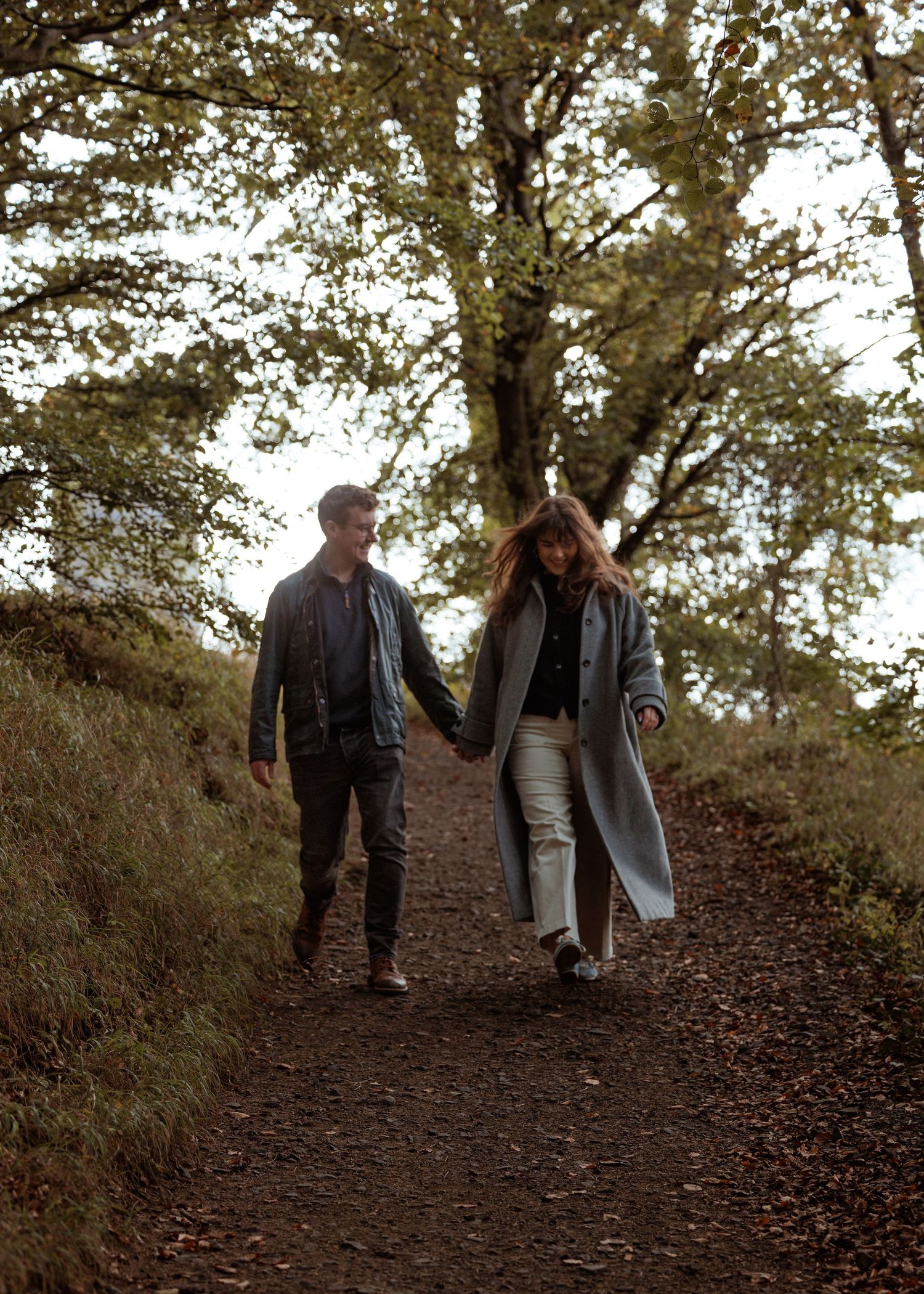 A couple takes a romantic autumn stroll along a tree-lined dirt path in nature.