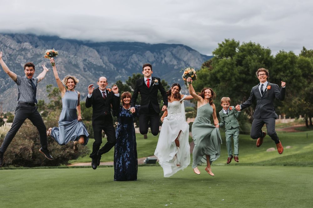 Wedding party members leap joyfully in synchronized poses on a lush golf course with mountains in the background.