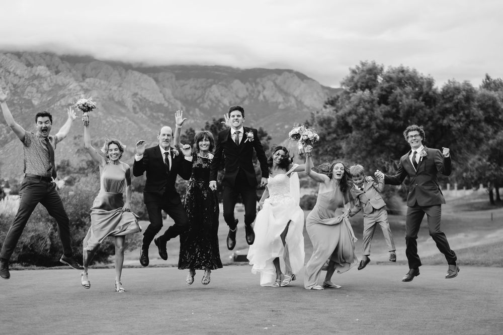 Black and white photo of wedding party jumping and dancing together outdoors with mountains in background.