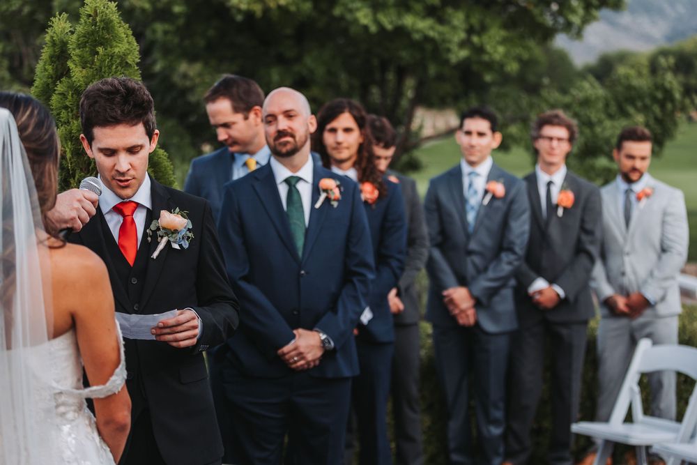 Wedding party in formal attire stands together outdoors in front of white chairs during garden ceremony.