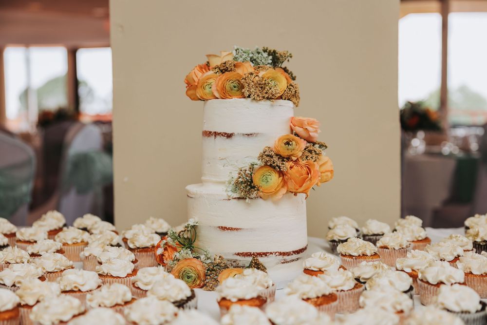 White wedding cake and cupcakes decorated with peach and orange flowers arranged on a dessert table at reception.