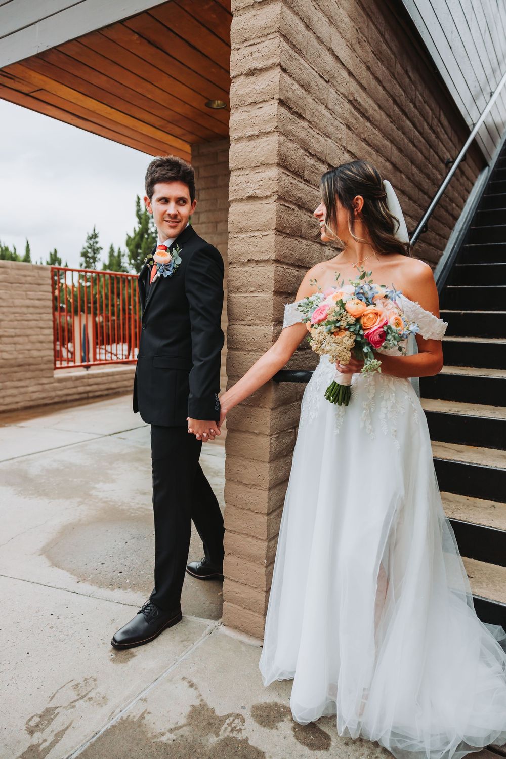 A couple in wedding attire shares intimate moments on outdoor stairs with brick walls and a beautiful floral bouquet.